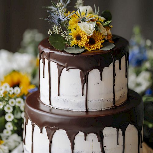 A two-tier cake with white frosting and chocolate drizzle, topped with yellow and white flowers, is placed on a white cake stand.