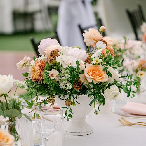 A table setting featuring a floral centerpiece with peach and white flowers, surrounded by glassware and pink napkins in a formal event setting.