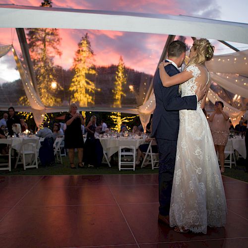 A couple dances under a tent at an outdoor event, surrounded by guests seated at tables with a stunning sunset and lit trees in the background.