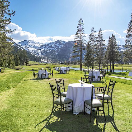 Tables and chairs set up on a grassy field with mountains in the background, under a clear blue sky and surrounded by tall trees.