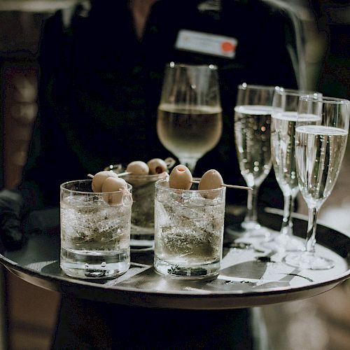 A person is holding a tray with three cocktails garnished with olives, a glass of white wine, and three glasses of champagne.