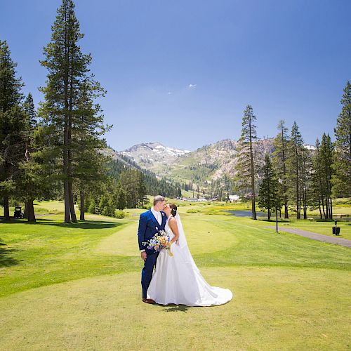 A couple in wedding attire is standing on grass, surrounded by trees and mountains, sharing a kiss in a serene outdoor setting.