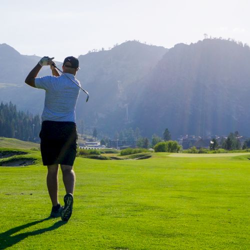 A person is playing golf on a lush green course surrounded by mountains and trees, with sunlight casting shadows on the landscape.