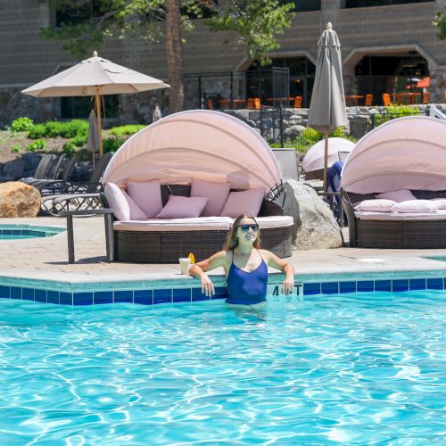 A woman lounges by a sunny outdoor pool at a resort, with white umbrellas and cushioned beds in the background.