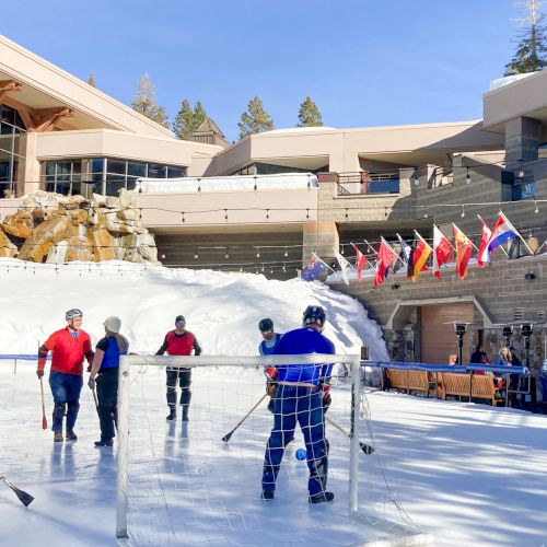 People are playing hockey on an outdoor ice rink with buildings and flags in the background, under a clear sky.