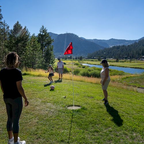 A sunny mountain meadow with people walking on the grass toward a red-flag marker by a lake for outdoor activities.