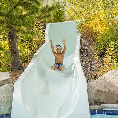 A joyful man slides down a white water slide through a sunny outdoor garden.