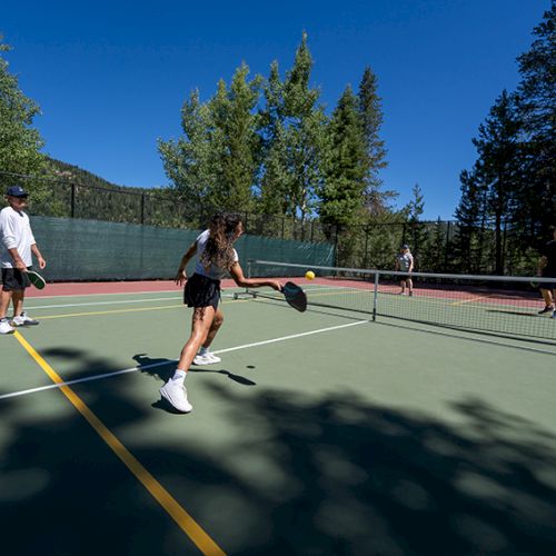 People play pickleball on a sunny outdoor court, with one woman swinging a paddle as others watch.