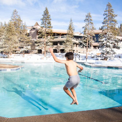 A shirtless man jumps into an outdoor heated pool at a snowy mountain resort, with pine trees and buildings behind him.