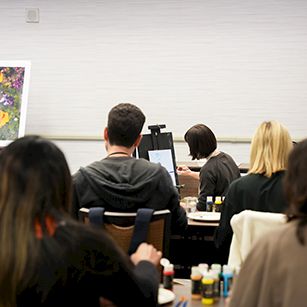 Art class: students seated at desks, facing an instructor, with an easel painting on the left.