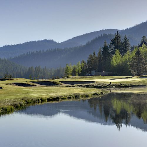 A tranquil golf course near a reflective lake, surrounded by lush greenery and distant mountains under a clear blue sky, creating a serene atmosphere.