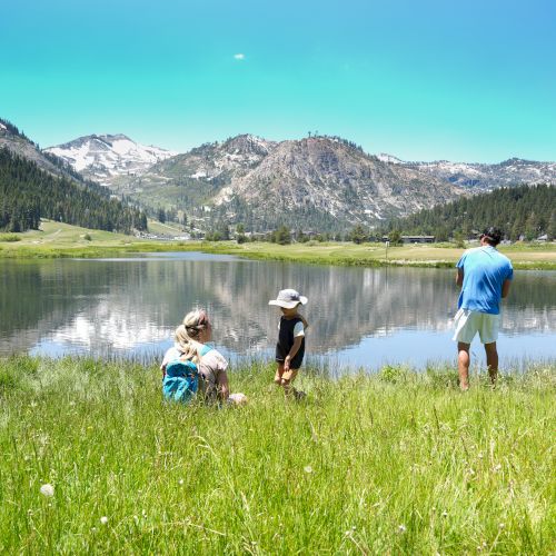 Three people enjoy a serene lakeside setting with mountainous views; one fishing, two seated on the grass, with a chair on the left.