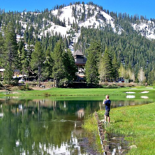 A person stands by a serene lake surrounded by lush greenery and snowy mountain peaks in the background, capturing the scenic view.