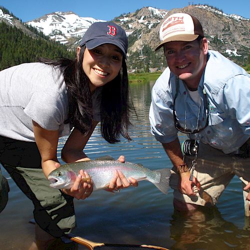 Two people are standing in a lake holding a fish, with mountains and trees in the background.