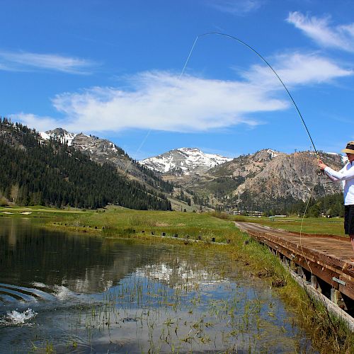 A person is fishing from a wooden dock in a serene mountain landscape with a calm lake and snowy peaks under a clear, blue sky.