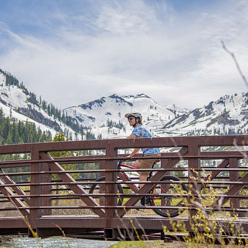 A person rides a bicycle on a wooden bridge with snow-capped mountains in the background and greenery around. The sky is partly cloudy.