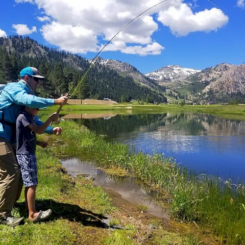 Two people are fishing beside a scenic lake with mountains, trees, and a blue sky in the background.