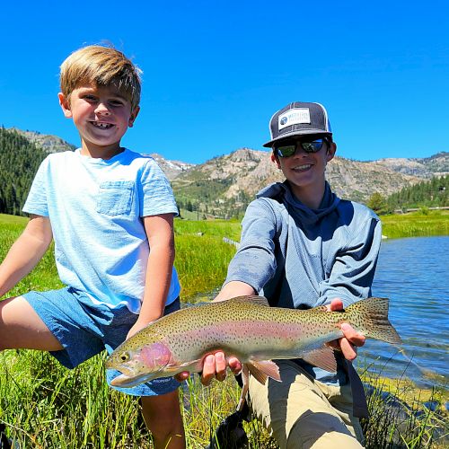 A boy and a man are outdoors by a lake holding a large fish, smiling, with mountains and blue skies in the background.