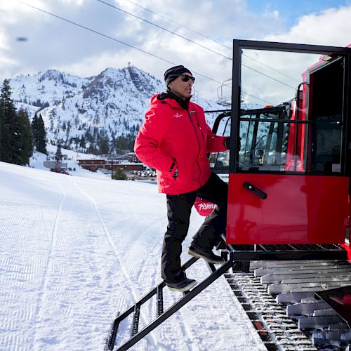 A person dressed in red steps into a large red snowcat vehicle on a snowy mountain with pine trees and a lodge in the background.