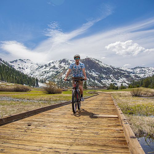 A person is riding a bike on a wooden pathway through a scenic landscape featuring snow-capped mountains and a clear blue sky.