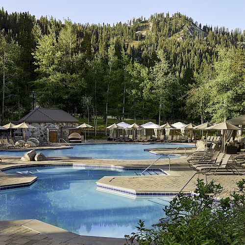 An outdoor resort pool area with lounge chairs and umbrellas, surrounded by trees and mountains in the background.