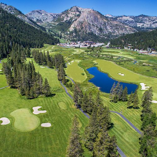 An aerial view of a lush golf course surrounded by trees, with a large pond, mountains in the background, and scattered buildings.