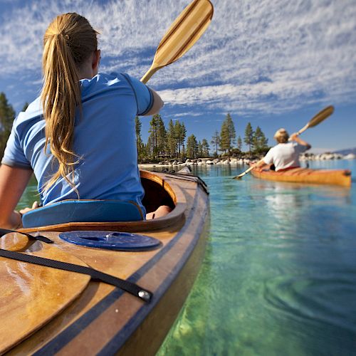 Two people are kayaking on a calm, clear body of water with a forest and mountains in the background. The sky is partly cloudy.