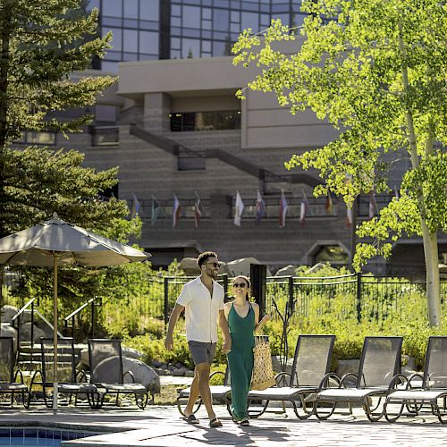 A couple is walking beside a swimming pool, surrounded by lounge chairs and greenery, with a modern building in the background.