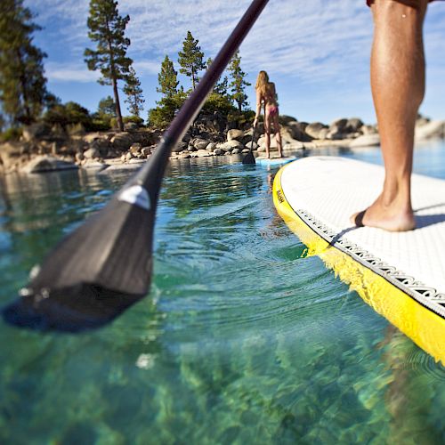 Two people are enjoying paddleboarding on clear, calm water with a scenic background of trees and rocks under a blue sky.