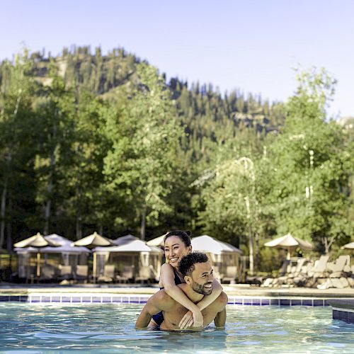 A couple is enjoying themselves in a pool surrounded by lounge chairs, umbrellas, and a scenic forested mountain backdrop.