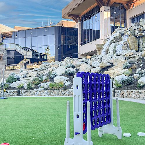 Large outdoor Connect Four game on a grassy area in front of a modern building with glass windows and rocky landscaping in the background.