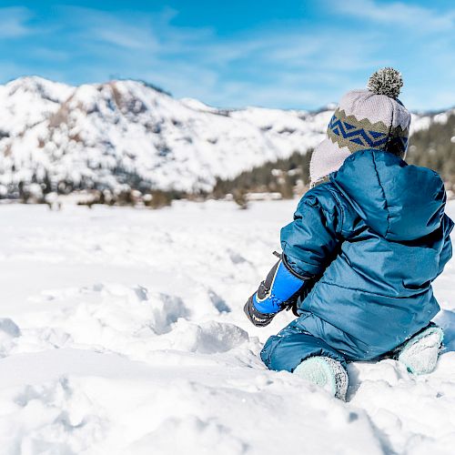 A child in winter clothing sits in the snow with a mountainous background under a clear blue sky.