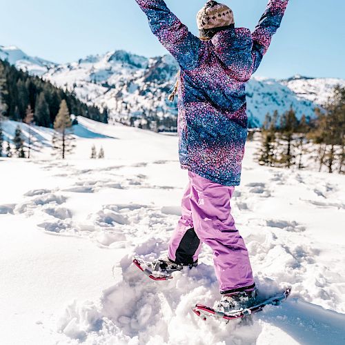 A person wearing colorful winter clothing and snowshoes stands in a snowy landscape with arms raised, enjoying the mountain scenery under a clear sky.