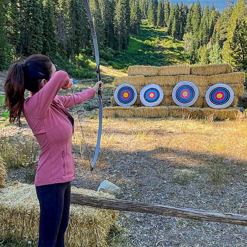 A person is practicing archery outdoors, aiming a bow at four circular targets lined up against hay bales, with a forest background.