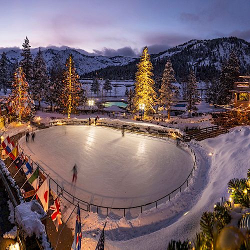 A snow-covered scene with a lit ice skating rink, decorated trees, and surrounding buildings against a mountain backdrop at twilight.