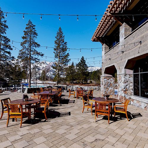 Outdoor patio with wooden tables and chairs, string lights overhead, trees, and mountains in the background, beside a stone building.