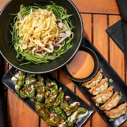 A bowl of shredded vegetables and egg, fried dumplings with dipping sauce, and grilled peppers on a wooden table with black napkins.