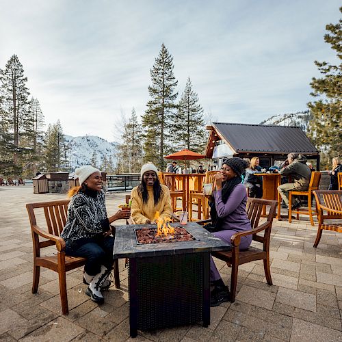 Three people sitting around a fire pit on a patio, surrounded by snow-covered mountains and trees, with a small bar structure in the background.