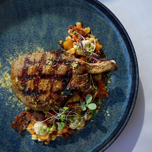 A grilled pork chop served on a blue plate with a bed of vegetables and microgreens garnished on top, beautifully plated.