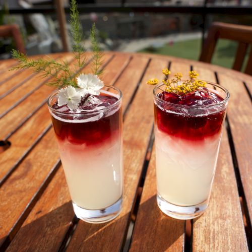 Two tall drinks on a wooden patio table&mdash;red layered beverages in glasses, garnished with edible flowers and herbs.