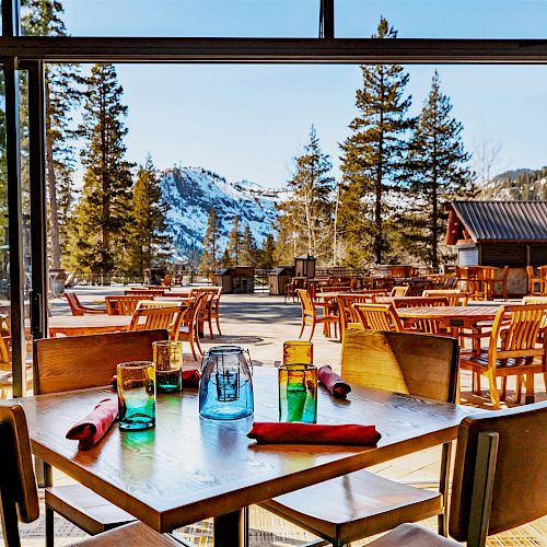 A wooden table with colorful glassware and napkins set in a restaurant with a stunning outdoor view of mountains and trees on a sunny day.