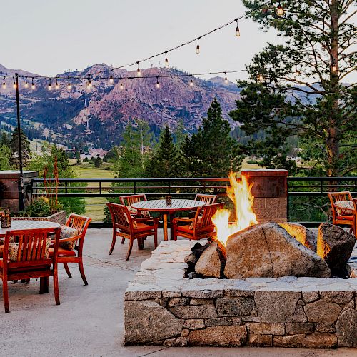 An outdoor patio with a stone fire pit, surrounded by wooden chairs and tables, overlooks mountains and trees with string lights above.