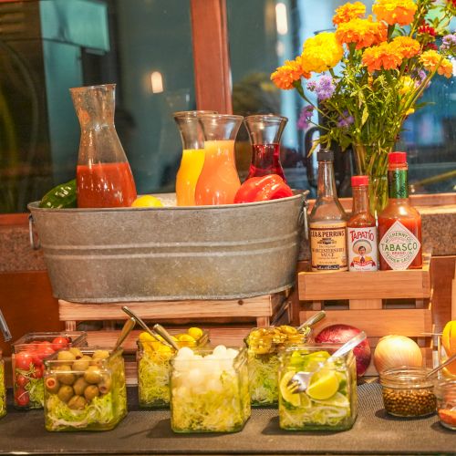 A brunch drink bar setup with jars of juices, condiments for Bloody Mary/Mimosa, and colorful flowers on display, inviting you to mix your own,