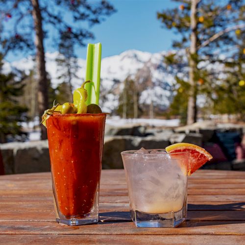 Two tall cocktails with green garnish and grapefruit slices sit on a wooden patio table with snowy mountains in the background.