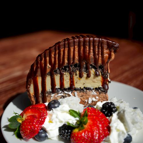 Slice of cheesecake topped with chocolate syrup, served with whipped cream and fresh strawberries, blueberries, and blackberries.