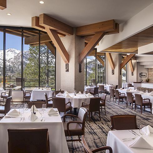 A stylish restaurant dining room with set tables, large windows, and a stone fireplace, overlooking snowy mountains through the glass.