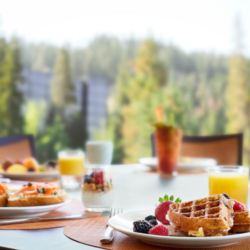 The image shows a table with breakfast foods including waffles, fruit, yogurt, orange juice, pastries, and coffee, set outdoors with a scenic view.