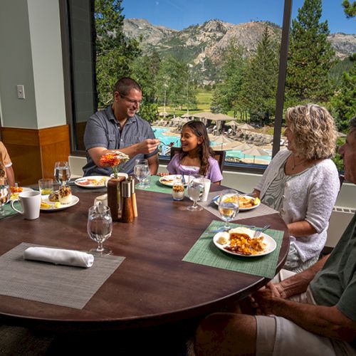 A family shares a meal at a window table, with drinks and plates while mountains and greenery stretch outside.