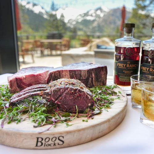 A wooden board with raw steaks, bottles of Frey Ranch whiskey, two glasses of whiskey, and a scenic mountain view in the background.