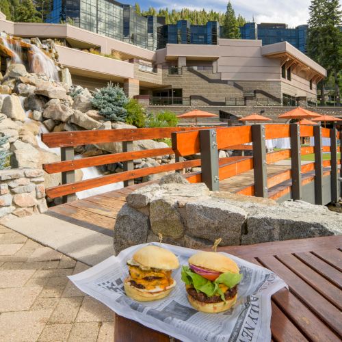 Outdoor patio by a rocky waterfall, burgers on a table and orange umbrellas, with a modern lodge and pine trees in view.